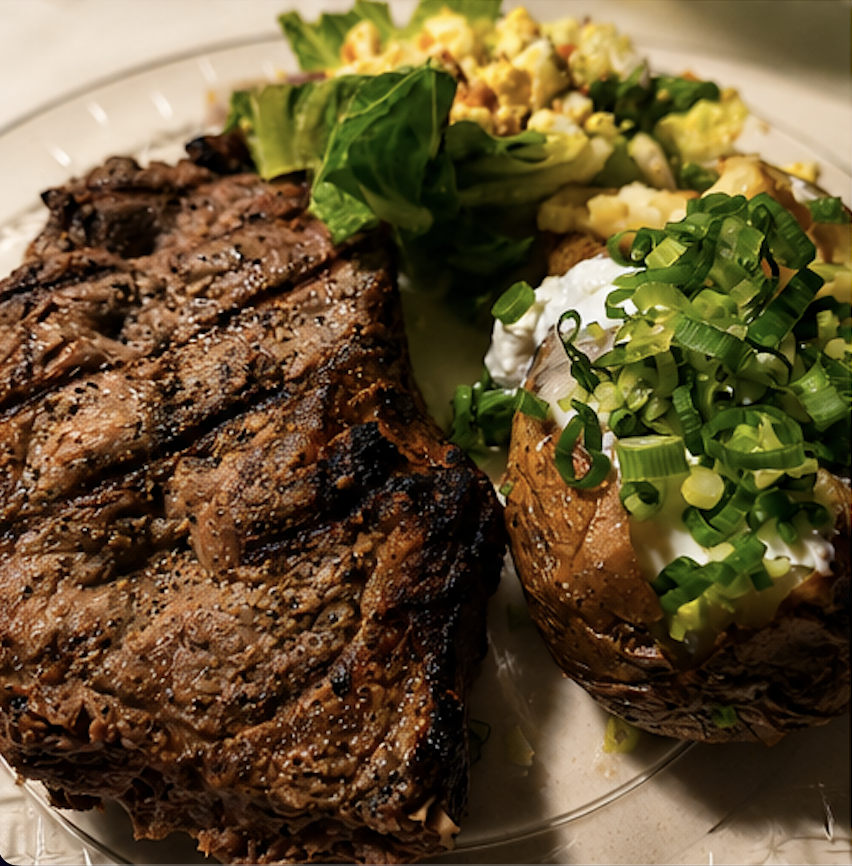Grilled ribeye steak plated with a loaded baked potato topped with sour cream and scallions, and a fresh side salad